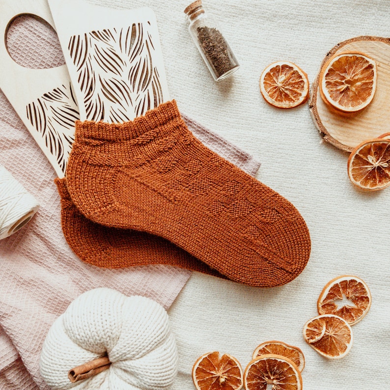 May include: A pair of rust-colored knitted ankle socks with a textured pattern. The socks are displayed on a light-colored surface, surrounded by decorative elements including dried orange slices, a wooden cutout, and a small glass bottle.