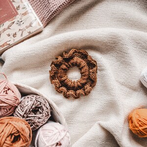 May include: A brown knitted hair scrunchie with a ruffled edge, surrounded by balls of yarn in various shades of pink, brown, and orange.