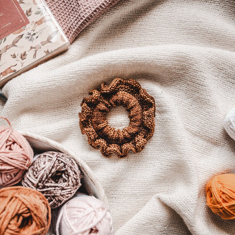 May include: A brown knitted hair scrunchie with a ruffled edge, surrounded by balls of yarn in various shades of pink, brown, and orange.