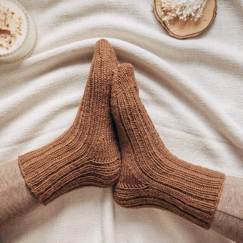 May include: A pair of brown knitted socks with a ribbed texture. The socks are folded over and placed on a white fabric background.