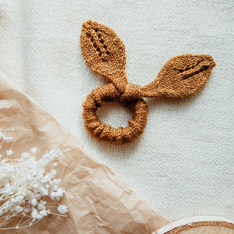 May include: A brown knitted hair scrunchie with two leaf-shaped ends. The scrunchie is tied in a knot and the ends are spread out.