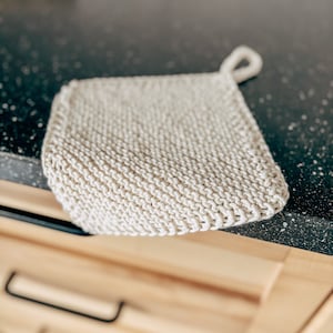 May include: A close-up of a white knitted dishcloth with a loop handle. The dishcloth is lying on a black countertop.