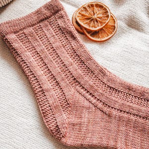 May include: A close-up of a pink knitted sock with a ribbed texture. The sock is lying on a white surface.