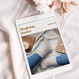 May include: A tablet displays an image of "Seafoam Socks" with a pair of light gray knitted socks. The socks are worn with blue jeans. The tablet is on a white surface with a floral arrangement in the background.
