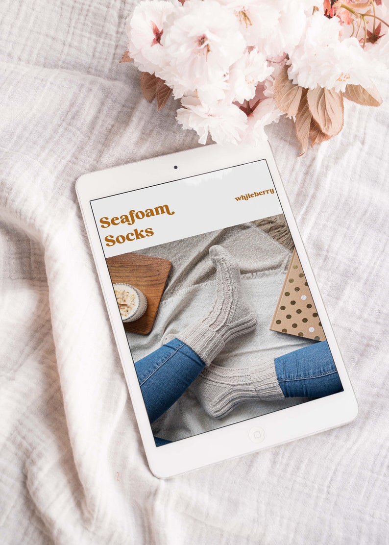 May include: A tablet displays an image of "Seafoam Socks" with a pair of light gray knitted socks. The socks are worn with blue jeans. The tablet is on a white surface with a floral arrangement in the background.
