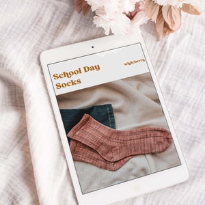 May include: A white tablet displaying a digital download for a knitting pattern called "School Day Socks" by whileberry. The pattern features a pair of pink socks on a fabric background.
