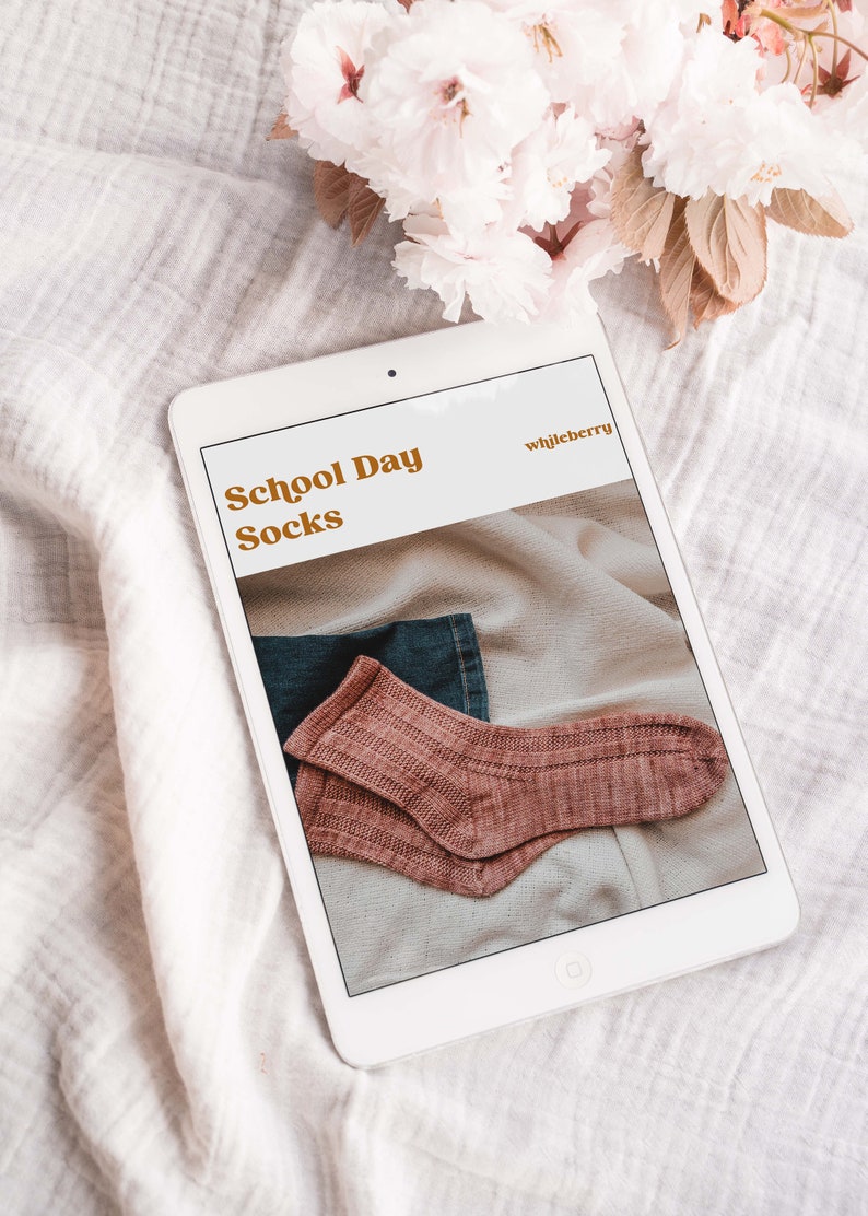 May include: A white tablet displaying a digital download for a knitting pattern called "School Day Socks" by whileberry. The pattern features a pair of pink socks on a fabric background.
