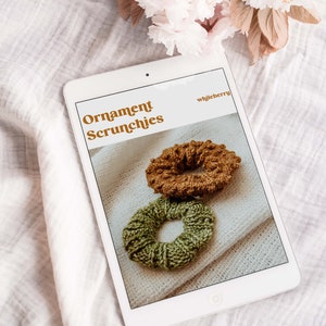 K&ouml;nnte beinhalten: Ein digitales Tablet zeigt eine Website mit dem Text "Ornament Scrunchies" und dem Website-Namen "whileberry". Die Website zeigt zwei gestrickte Haargummis, eines braun und eines gr&uuml;n, auf wei&szlig;em Hintergrund.