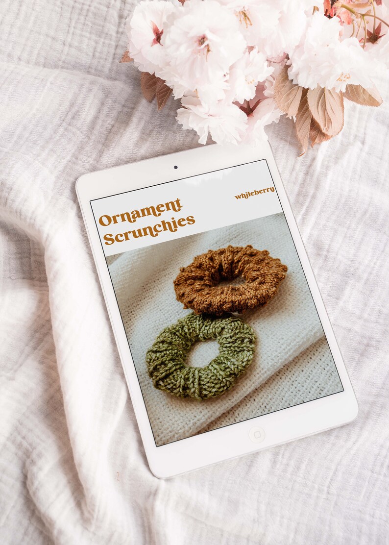 K&ouml;nnte beinhalten: Ein digitales Tablet zeigt eine Website mit dem Text "Ornament Scrunchies" und dem Website-Namen "whileberry". Die Website zeigt zwei gestrickte Haargummis, eines braun und eines gr&uuml;n, auf wei&szlig;em Hintergrund.