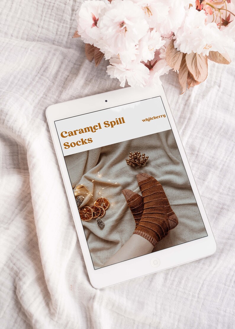Puede incluir: Una tableta digital que muestra un patr&oacute;n de tejido para "Caramel Spill Socks" de "whileberry". El patr&oacute;n presenta un primer plano de calcetines rayados marrones sobre un fondo blanco con rodajas de naranja secas y pi&ntilde;as.