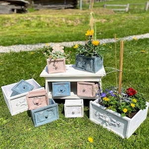 Könnte beinhalten: Ein Set aus sieben weißen, rosa und blauen bemalten Holzkasten-Pflanzgefäßen mit Blumendesigns. Die Kästen sind auf einem weißen Holztisch und auf dem Gras angeordnet. Einige der Kästen sind mit Pflanzen und Blumen gefüllt.