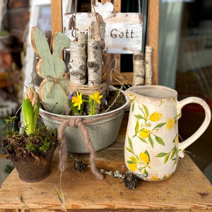 May include: A rustic wooden table displays a spring-themed arrangement. It features a ceramic pitcher with lemon designs, a metal planter with daffodils, a wooden bunny, and a small potted plant. The background includes a wooden chair with a sign that says "Gott".