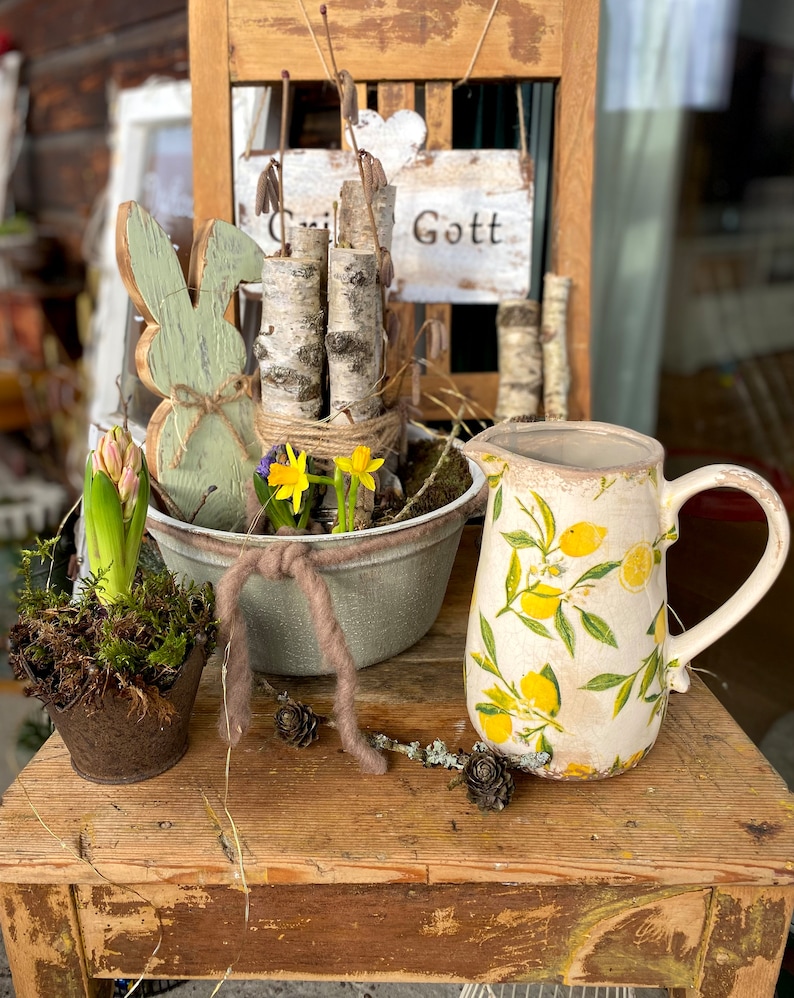 May include: A rustic wooden table displays a spring-themed arrangement. It features a ceramic pitcher with lemon designs, a metal planter with daffodils, a wooden bunny, and a small potted plant. The background includes a wooden chair with a sign that says "Gott".
