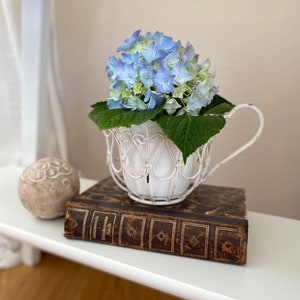 Könnte beinhalten: Ein weißer Draht-Teetassen-Pflanztopf gefüllt mit blauen und grünen Hortensien und grünen Blättern, der auf einem alten Buch steht. Links befindet sich eine dekorative Kugel. Die Teetasse hat einen Henkel und herzförmige Ausschnitte.