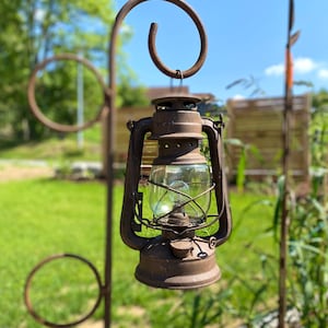 Könnte beinhalten: Eine verwitterte, rostige Metalllaterne mit einer klaren Glaskugel hängt an einem dekorativen, gebogenen Metallhaken. Die Laterne hängt im Freien vor dem Hintergrund von grünem Gras und einem strahlend blauen Himmel, was auf eine Garteneinstellung hindeutet.