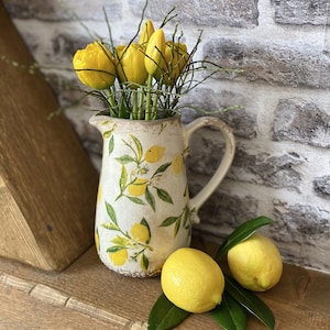 May include: A white ceramic pitcher with a lemon design holds yellow tulips and twigs. Two fresh lemons and green leaves rest on a wooden surface. The pitcher has a handle and a slightly distressed finish, evoking a rustic aesthetic.