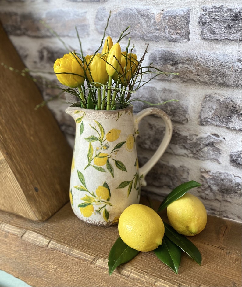 May include: A white ceramic pitcher with a lemon design holds yellow tulips and twigs. Two fresh lemons and green leaves rest on a wooden surface. The pitcher has a handle and a slightly distressed finish, evoking a rustic aesthetic.