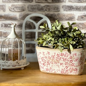 Peut inclure: Un pot de fleurs en céramique blanche avec un motif floral rouge contenant une plante verte, placé à côté d'un bougeoir en métal blanc en forme de cage à oiseaux avec une bougie rose, le tout posé sur une surface en bois.