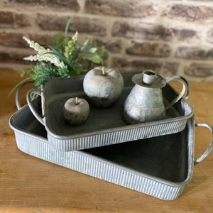 Könnte beinhalten: Zwei graue Metalltabletts mit geriffelten Kanten und Griffen. Das obere Tablett hat einen kleinen grauen Keramikapfel und einen grauen Keramikkannen. Das untere Tablett ist größer und steht auf einer Holzoberfläche.