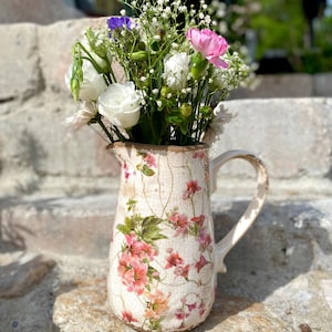 Könnte beinhalten: Eine cremefarbene Kanne im Vintage-Stil mit floralem Design, gefüllt mit einem Strauß frischer Blumen. Die Blumen umfassen weiße Rosen, rosa Nelken und kleine weiße und violette Blüten. Die Kanne hat einen Henkel und steht auf einer Steinoberfläche.