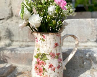 May include: A vintage-style cream-colored pitcher with a floral design, filled with a bouquet of fresh flowers. The flowers include white roses, pink carnations, and small white and purple blossoms. The pitcher has a handle and is placed on a stone surface.