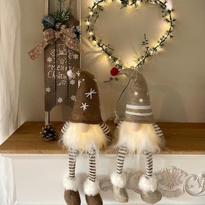 May include: Two plush gnome figurines with long white beards and dangling striped legs. One gnome wears a brown hat with white snowflakes, the other a tan hat with white dots. Both feature internal warm white LED lights and soft fabric textures.