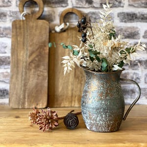 Könnte beinhalten: Ein Stillleben mit einer verwitterten Metallkanne, gefüllt mit getrockneten Blumen und Grün. Zwei Holzschneidebretter mit Ringgriffen befinden sich im Hintergrund. Dekorative Trockenblumen und ein Tannenzapfen im Vordergrund.