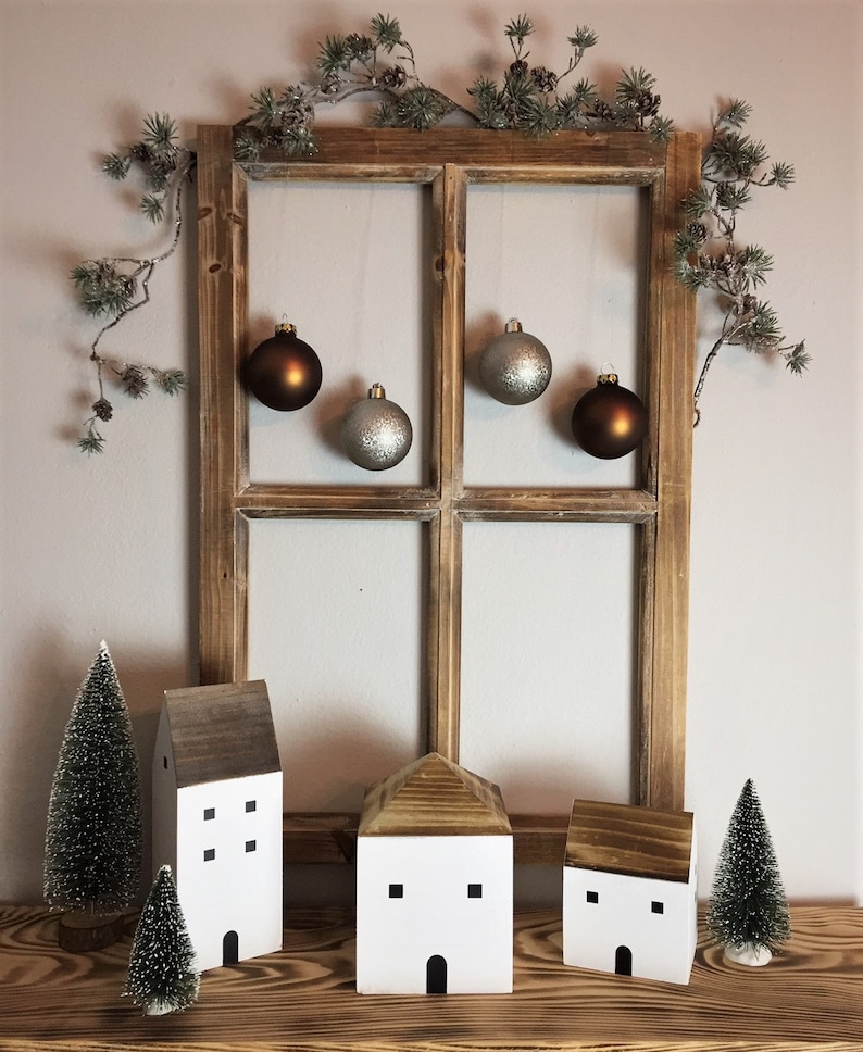 May include: A rustic wooden window frame decorated with a pine garland and four hanging ornaments in matte brown and glittery silver. Below are three white wooden house figurines with dark roofs and two small green bottle brush pine trees.