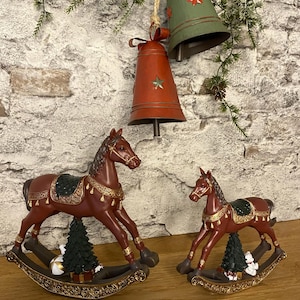 Könnte beinhalten: Zwei dekorative Schaukelpferde-Figuren im Weihnachtsthema. Die Pferde sind braun mit goldenen Akzenten und grünen Satteldecken. Ein kleiner Weihnachtsbaum und eine weiße Verzierung befinden sich auf der Wippenbasis. Rote und grüne Glocken hängen darüber.
