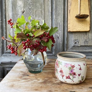 Peut inclure: Un vase en verre transparent contient un arrangement de baies rouges et de feuilles vertes. À côté, un petit pot en céramique est orné d'un motif floral rose et violet. Le tout est posé sur une surface en bois rustique.