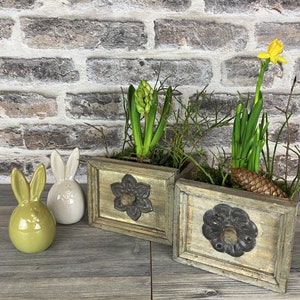 Könnte beinhalten: Zwei Holzpflanzkästen mit floralen Metallakzenten, gefüllt mit Grün und Frühlingsblumen, darunter eine gelbe Narzisse. Zwei Keramik-Hasenfiguren in Grün und Weiß stehen links. Der Hintergrund ist eine Backsteinmauer.