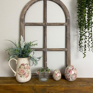 May include: A decorative arched wooden window frame with a dark brown finish. A cream-colored ceramic pitcher with a pink rose floral pattern holds green foliage. A small ribbed glass jar and two egg-shaped ornaments with floral prints are present.