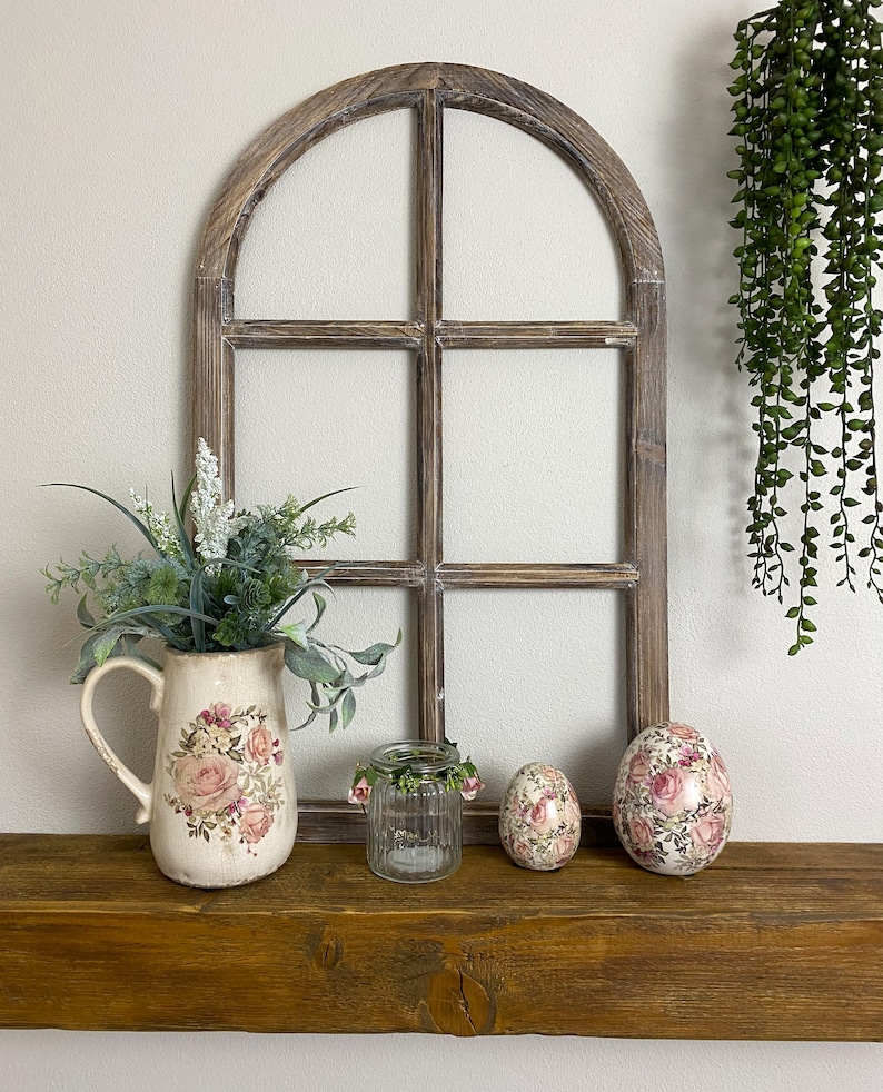 May include: A decorative arched wooden window frame with a dark brown finish. A cream-colored ceramic pitcher with a pink rose floral pattern holds green foliage. A small ribbed glass jar and two egg-shaped ornaments with floral prints are present.