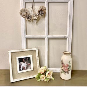 May include: A white distressed wooden window frame with a hanging floral ornament, a square picture frame containing a photograph of a cat, a small bouquet of light pink artificial flowers, and a cylindrical vase with a floral decal.