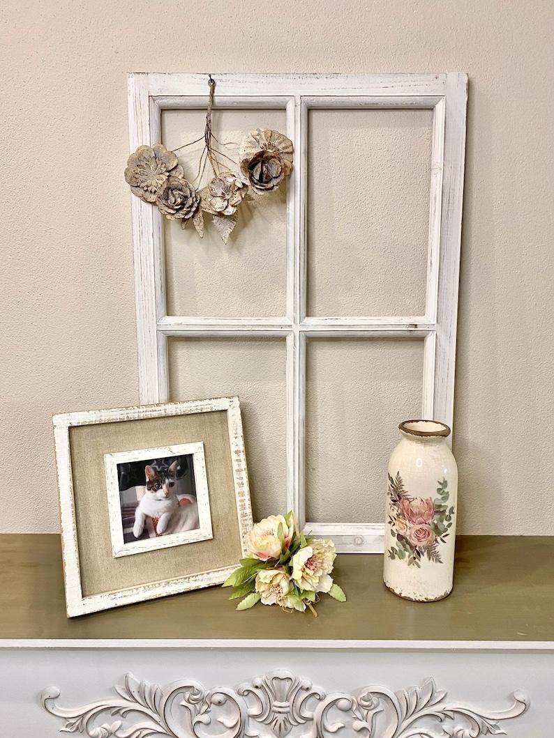 May include: A white distressed wooden window frame with a hanging floral ornament, a square picture frame containing a photograph of a cat, a small bouquet of light pink artificial flowers, and a cylindrical vase with a floral decal.