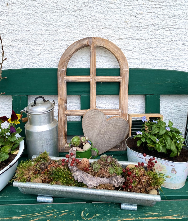 May include: A decorative wooden arched window frame, a wooden heart, a silver metal milk can, a green enamel mug with succulents, and a rectangular galvanized metal planter filled with moss, red berries, and succulents on a green bench.