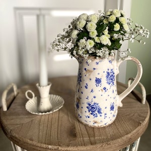 Peut inclure: Une cruche crème avec des motifs floraux bleus, remplie de fleurs blanches, est posée sur un plateau rond en bois. Une bougie et un bougeoir blancs sont également sur le plateau. Le plateau est sur un support en métal blanc.