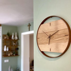 Può includere: Un orologio da parete rotondo con un quadrante in legno chiaro e lancette e indicatori marrone scuro. Il quadrante è racchiuso in un anello di metallo marrone scuro. L'orologio è montato su una parete verde chiaro.