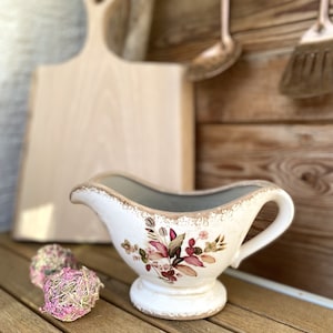 Może przedstawiać: Biała ceramiczna sosjerka z kwiatowym wzorem w odcieniach r&oacute;żowego, zielonego i brązowego. Sosjerka ma postarzaną powierzchnię i uchwyt po prawej stronie.