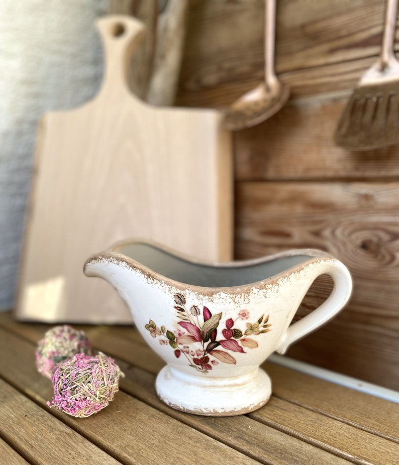 Może przedstawiać: Biała ceramiczna sosjerka z kwiatowym wzorem w odcieniach r&oacute;żowego, zielonego i brązowego. Sosjerka ma postarzaną powierzchnię i uchwyt po prawej stronie.
