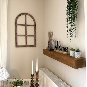 May include: A wooden arched window frame wall decor, a floating wooden shelf holding a black wire geometric candle holder, a black Home sign, and two small patterned pots with green plants, plus a hanging green plant and two wooden candlesticks.