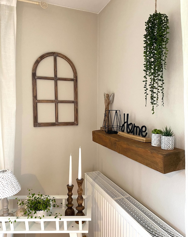 May include: A wooden arched window frame wall decor, a floating wooden shelf holding a black wire geometric candle holder, a black Home sign, and two small patterned pots with green plants, plus a hanging green plant and two wooden candlesticks.