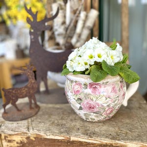 Könnte beinhalten: Ein Blumenarrangement in einem dekorativen Topf mit Henkel. Der Topf ist mit rosa Rosen und grünen Blättern verziert. Das Arrangement zeigt weiße Blüten mit gelben Zentren und grünes Laub. Im Hintergrund befinden sich rustikale Metall-Hirschfiguren.