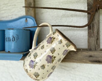 May include: A ceramic pitcher with a cream-colored base featuring a repeating pattern of purple and green hydrangea flowers and a visible crackle glaze finish. The pitcher has a curved handle and a spout, positioned at an angle in the frame.