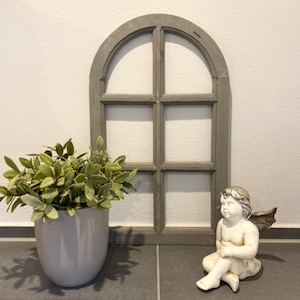 May include: A gray arched wooden window frame with six panes, a potted green plant in a smooth gray ceramic pot, and a small white and silver angel figurine with wings sitting in a relaxed pose on a dark gray tiled surface.