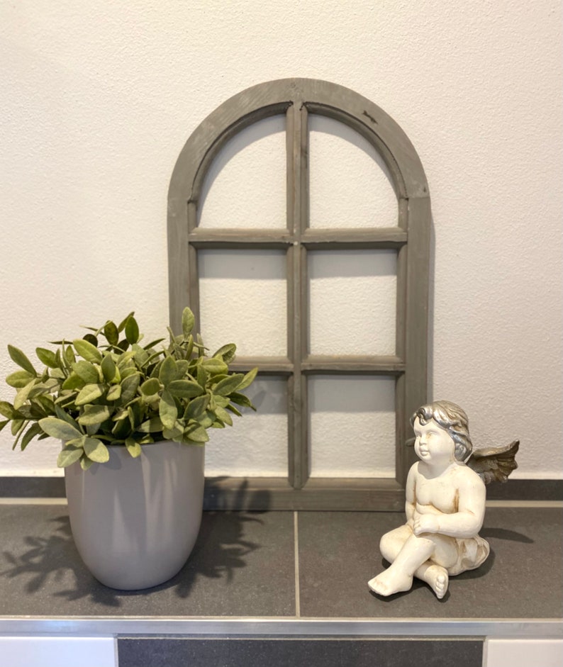 May include: A gray arched wooden window frame with six panes, a potted green plant in a smooth gray ceramic pot, and a small white and silver angel figurine with wings sitting in a relaxed pose on a dark gray tiled surface.
