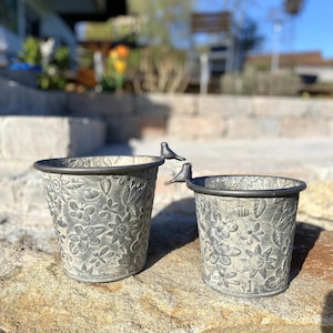 Può includere: Due vasi di fiori in metallo grigio con un design floreale e piccoli accenti di uccelli. I vasi sono appoggiati su una roccia grigia.