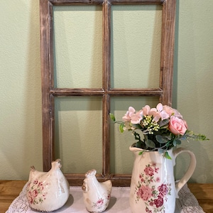 May include: A rectangular wooden window frame with a brown finish, two ceramic chicken figurines with pink floral patterns, and a ceramic pitcher featuring pink roses and green leaves, containing a bouquet of pink and white artificial flowers.