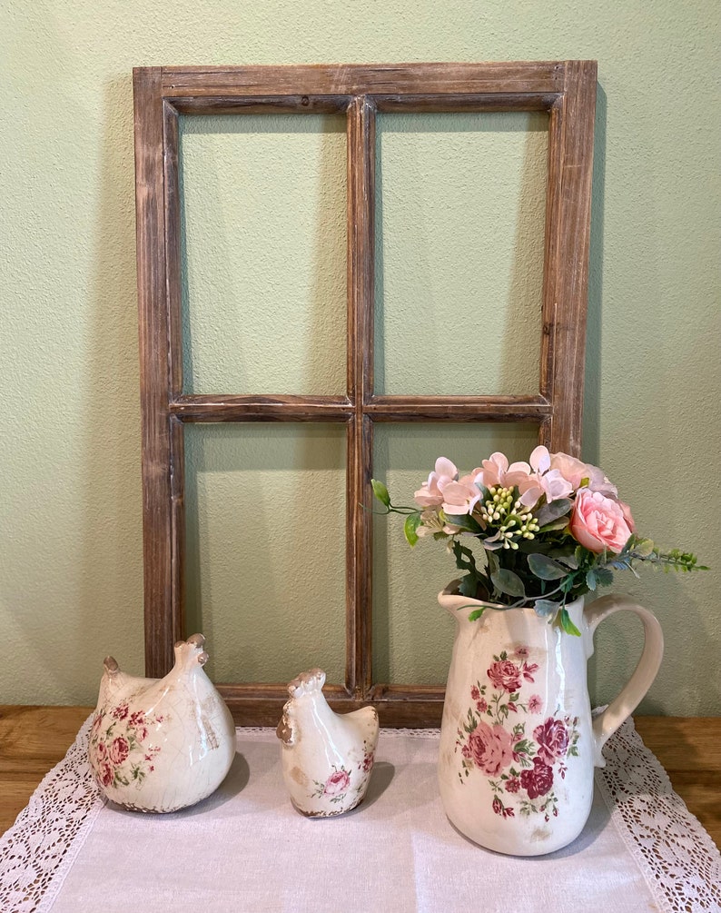 May include: A rectangular wooden window frame with a brown finish, two ceramic chicken figurines with pink floral patterns, and a ceramic pitcher featuring pink roses and green leaves, containing a bouquet of pink and white artificial flowers.