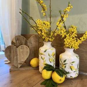 Könnte beinhalten: Zwei weiße Keramikvasen mit Zitronenmotiven halten leuchtend gelbe Forsythienzweige. Holzherzdekorationen und frische Zitronen mit grünen Blättern sind auf einer Holzoberfläche angeordnet und schaffen eine fröhliche, frühlingshafte Atmosphäre.